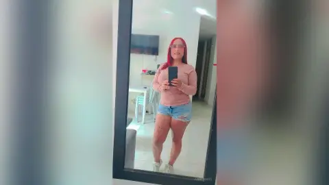 Red-haired woman in a pink long-sleeve top and denim shorts taking a casual mirror selfie in a bright room.