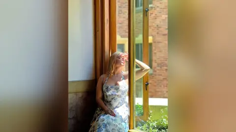 Blonde woman in a floral sundress sitting by a sunlit window in a bright, peaceful indoor setting.