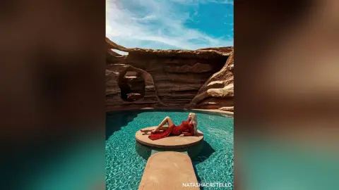 NatashaCastello relaxes in a red dress while lounging on a floating platform in a turquoise desert pool.