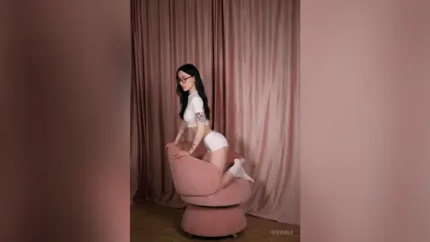 Young woman in white crop top and shorts sitting on a pink velvet chair against draped curtains.