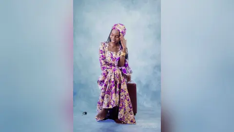 Smiling woman in a purple patterned African print maxi dress posing against a blue studio background.