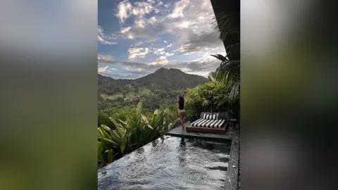 Woman in black swimwear standing by an infinity pool overlooking lush tropical mountains under a cloudy sky.