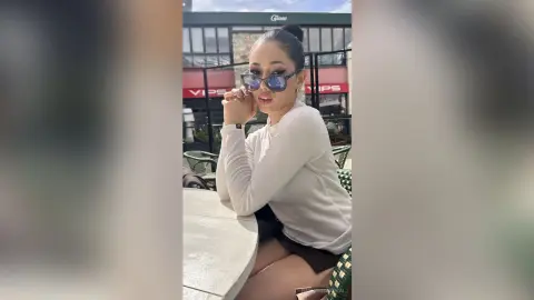 NashiroSakurai sits at a white table wearing a white long-sleeve top and dark skirt, looking at the camera through blue-tinted sunglasses.