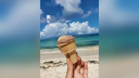 Hand holding a chocolate ice cream cone on a sunny tropical beach with blue ocean waves and white clouds.