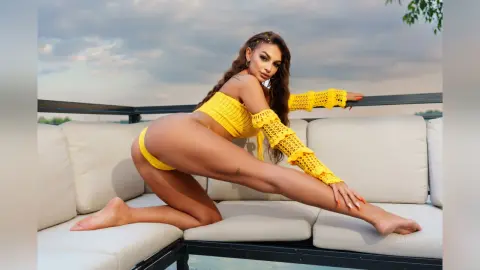 Anastasia poses naked on a white outdoor sofa wearing a bright yellow knit bra and matching bottom, showing her bare skin and legs.