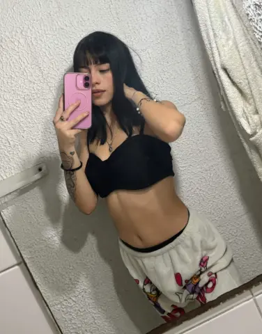 Young woman with black hair in a black crop top taking a casual mirror selfie in a bathroom.