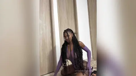 Young woman with long dark braids wearing a purple long-sleeve bodysuit in a minimalist indoor setting.
