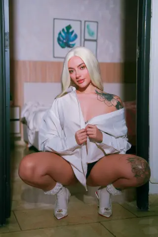 Blonde woman in an oversized white shirt squatting in a cozy bedroom setting with a relaxed mood.
