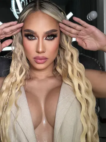 Glamorous woman with wavy blonde hair and dramatic eye makeup wearing a beige plunging neckline outfit.