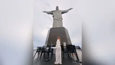 Woman in a white maxi dress standing before the Christ the Redeemer statue in Rio de Janeiro under a cloudy sky.