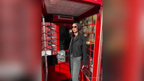 Stylish woman in a black jacket and sunglasses using a vintage red telephone booth in bright sunlight.