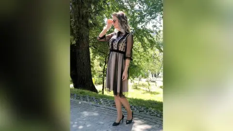 Elegant woman in a black sheer midi dress standing on a stone path in a sunny green park.