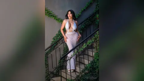 DayanaHille stands on a stone staircase wearing a white cut-out jumpsuit, posing against a grey wall with green ivy vines.