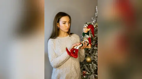 Woman in a white cable knit sweater holding a festive Christmas elf doll near a decorated silver tree.
