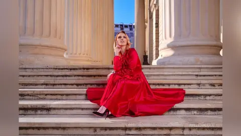 Elegant woman in a flowing red satin gown sitting on classical stone stairs against grand marble columns.