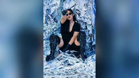 KimRoberth poses in a black bodysuit and sunglasses against a shiny silver foil background.