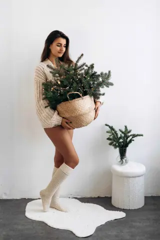 Young woman in cozy cream sweater and socks holding a small decorated Christmas tree in a bright minimalist room.