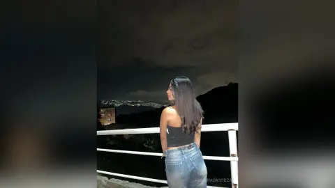 Young woman in a black tank top and blue jeans looking at city lights at night from a balcony.