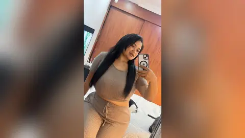 NiaCorelli poses in a mirror selfie wearing a tan ribbed two-piece crop top and matching lounge pants, showing her long dark hair and midriff.