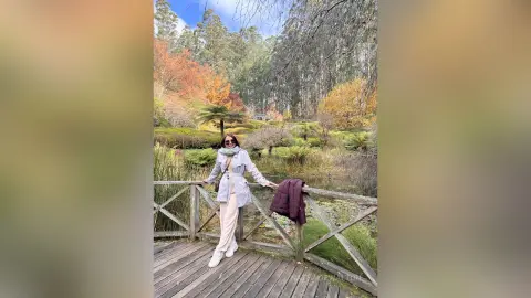 Woman in a grey trench coat and white trousers standing on a wooden pier by a scenic autumn lake forest.