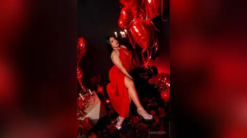 KimRoberth poses in a red slit dress and silver heels against a dark background with red heart balloons.
