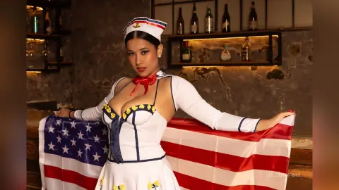 JuliahLewis poses in a white and blue sailor-style outfit while holding an American flag behind her.