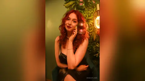 Redheaded woman in a black dress posing in a warm, moody indoor setting with lush green plants.