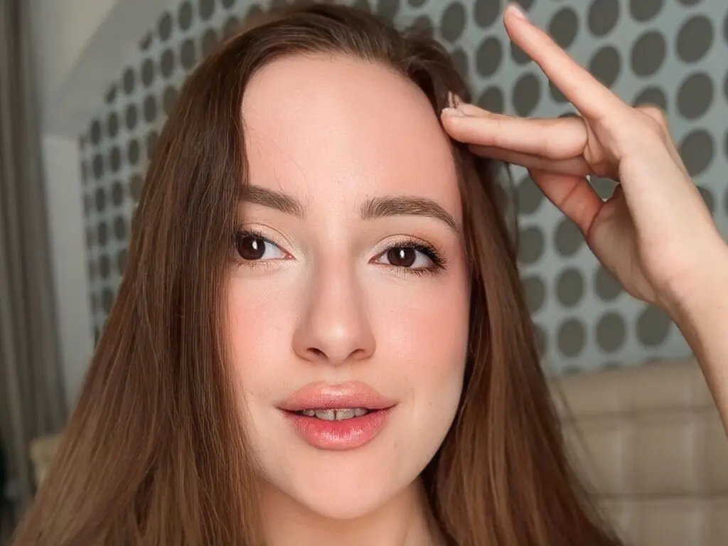 Young woman with long brown hair and natural makeup looking at camera in a bright indoor setting.