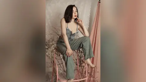 Young woman in denim overalls sitting on a pink chair in a dreamy, soft-lit studio setting.