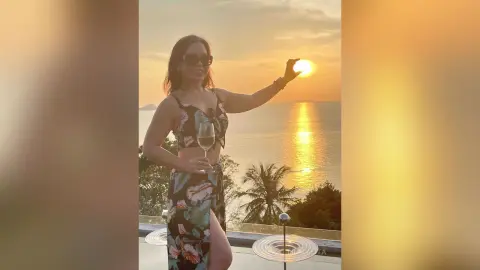 Smiling woman in floral maxi dress holding a wine glass against a golden sunset ocean backdrop.
