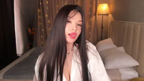 Young woman with long dark hair wearing a white shirt in a cozy, dimly lit bedroom setting.