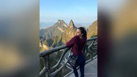 Smiling woman in a burgundy sweater and jeans standing on a scenic mountain lookout at sunset.
