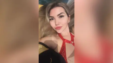 RaisaAllens poses in a red strap top with blonde hair, looking directly at the camera with a soft smile.