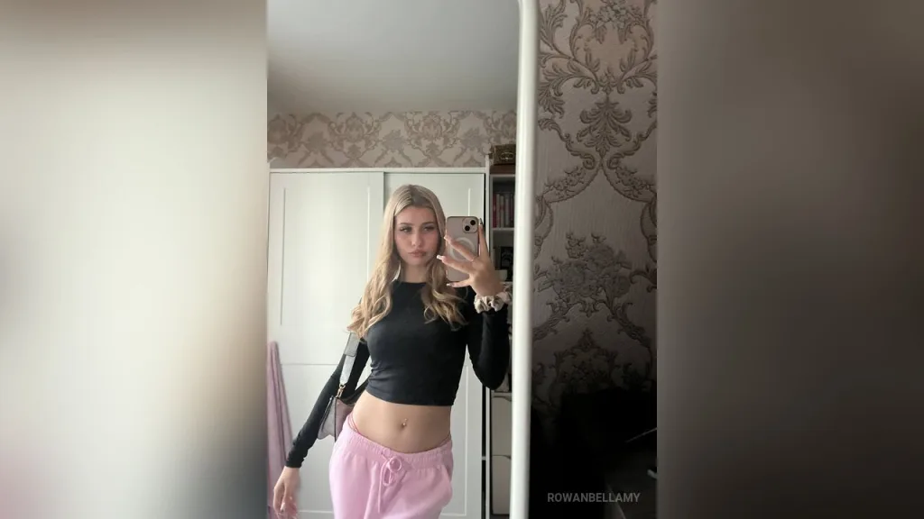 RowanBellamy poses with long blonde hair and light blue eyes, wearing a black top against a backdrop of brown curtains.