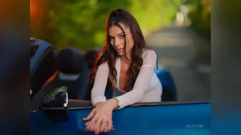 Elegant woman in a white long-sleeve top leaning on a blue convertible car during a golden hour sunset drive.