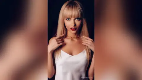 Blonde woman in a white satin top and gold heart necklace posing with a glamorous, moody expression.
