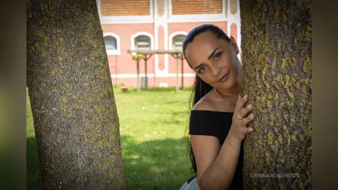Smiling woman in a black off-the-shoulder top peeking from behind a large tree in a sunny park.