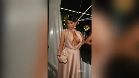 Dolce wears a tan halter-neck dress with a deep plunging neckline that shows her cleavage, holding a gold clutch bag.