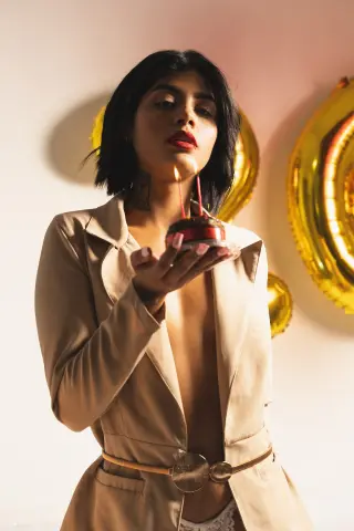 Elegant woman in a beige blazer holding a chocolate birthday cake with lit candles against a festive gold background.