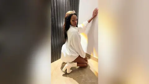 Elegant woman in a white oversized shirt and boots posing against a modern wall in soft, warm lighting.