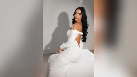 Alison poses in a white off-the-shoulder dress, showing her back and long black hair against a plain grey background.