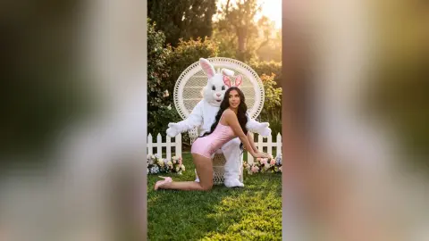 Woman in a pink bodysuit posing with a white bunny mascot in a sunny garden setting.