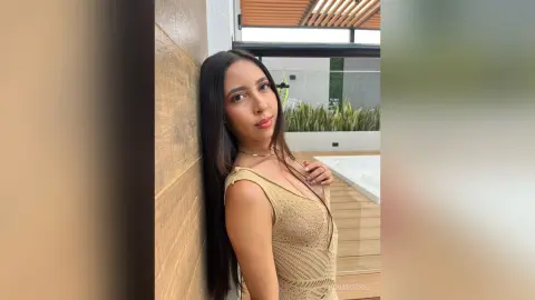 IrinaRussel with long black hair wearing a tan mesh dress, leaning against a wall outdoors.