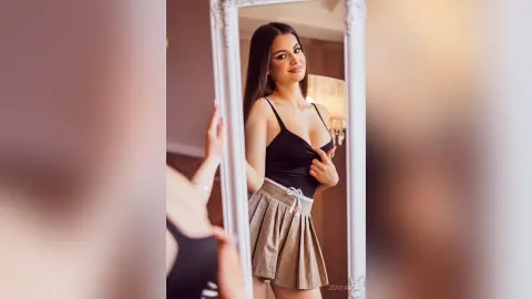 Young woman in a black bodysuit and tan skirt posing confidently in a mirror for a stylish indoor portrait.