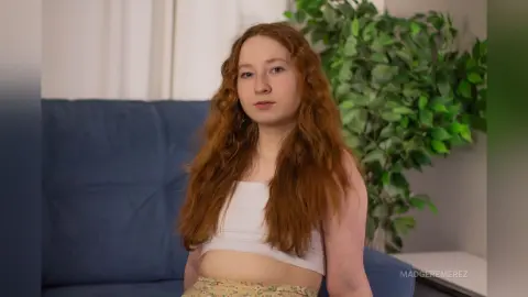Redheaded woman in a white crop top sitting on a blue sofa in a bright room with a green plant.