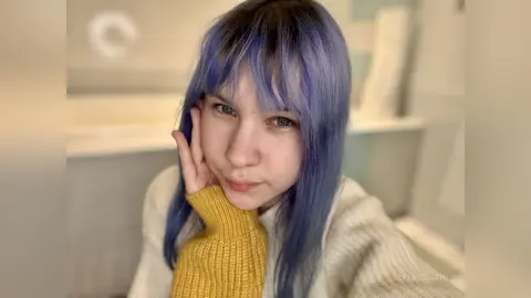 Young woman with purple hair wearing a cream sweater in a bright, minimalist indoor setting.