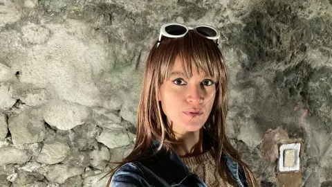 Young woman with brown bangs wearing sunglasses and a denim jacket against a rustic stone wall background.