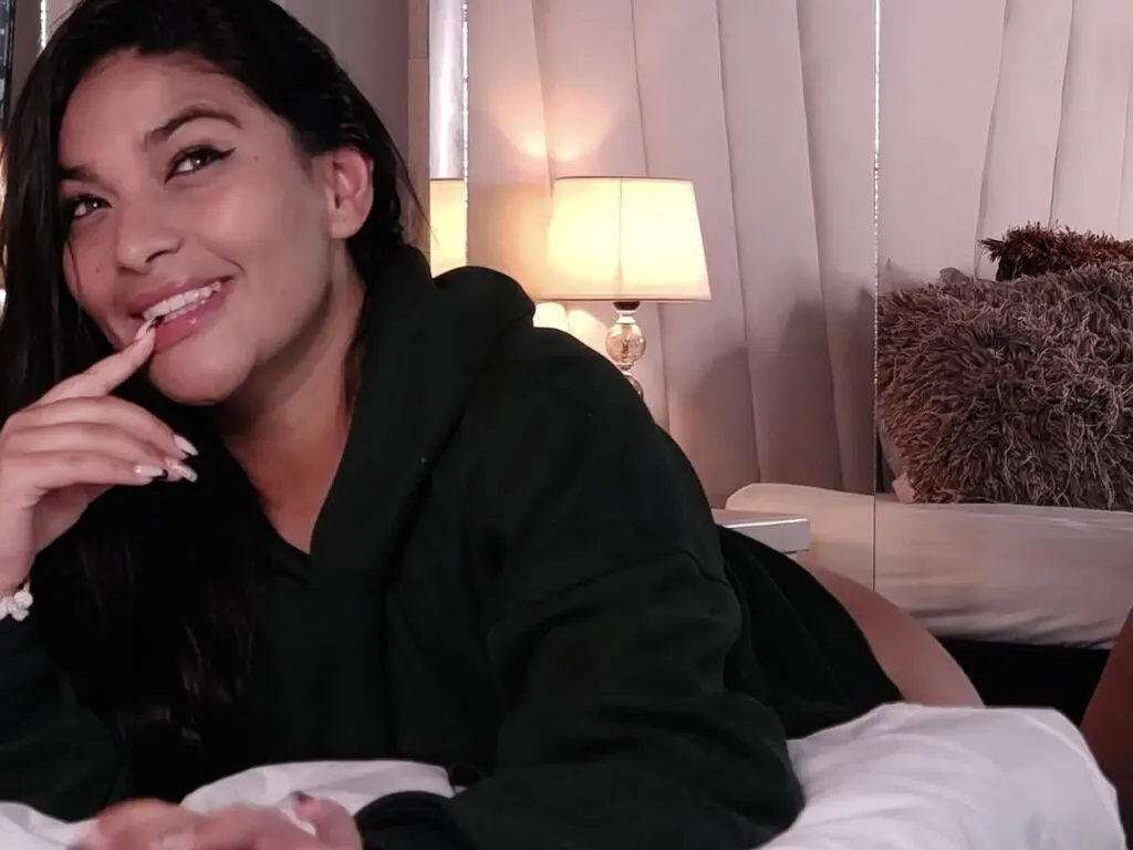 Smiling young woman in a cozy black hoodie relaxing on a white bed in a warm, dimly lit bedroom.