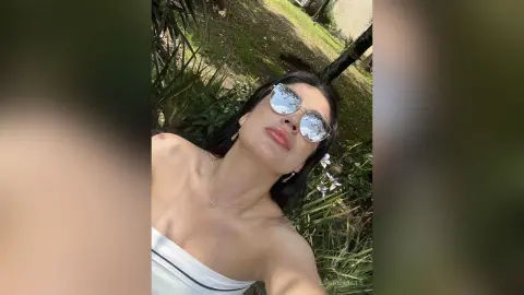 Young woman in sunglasses and white top smiling in a sunny outdoor garden setting.