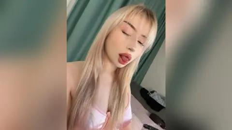 Blonde woman in a pink satin top posing playfully in a room with teal curtains.